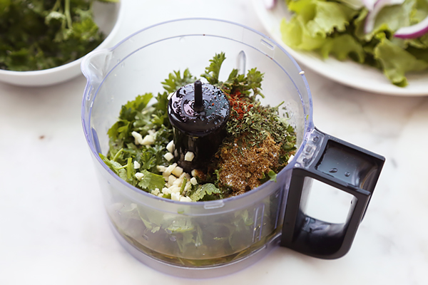 Cilantro, parsley, mint, garlic, and cumin loaded into a food processor bowl