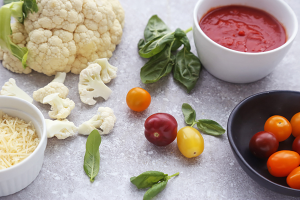 Whole cauliflower head, basil, multicolored cherry tomatoes, tomato sauce, and parmesan on a counter