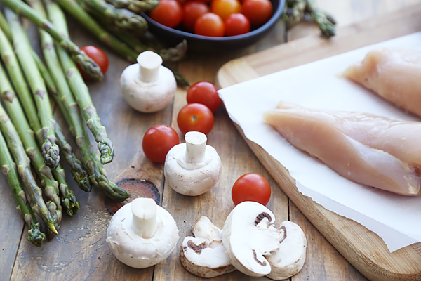 Whole chicken breasts, button mushrooms, asparagus spears, and cherry tomatoes on a cutting board