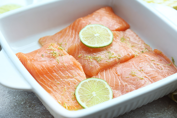 Three raw salmon fillets marinating with lime slices and zest in a white baking dish
