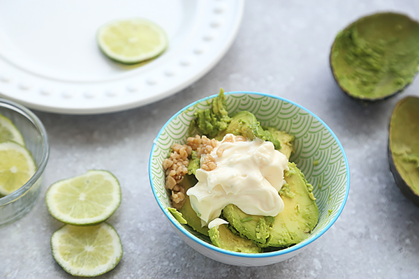Halved avocado, minced garlic, and mayonnaise in a small blue-rimmed bowl with limes nearby