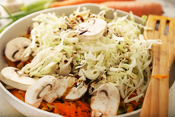 Shredded green cabbage and sliced button mushrooms piled over sauteed carrots in a frying pan