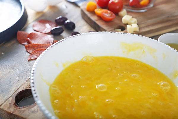 Whisked eggs in a white bowl with pepperoni, olives, tomatoes, and mozzarella cubes nearby