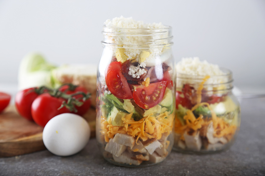 Cobb salad layered in mason jars with chicken, cheddar, avocado, tomato slices, and riced cauliflower