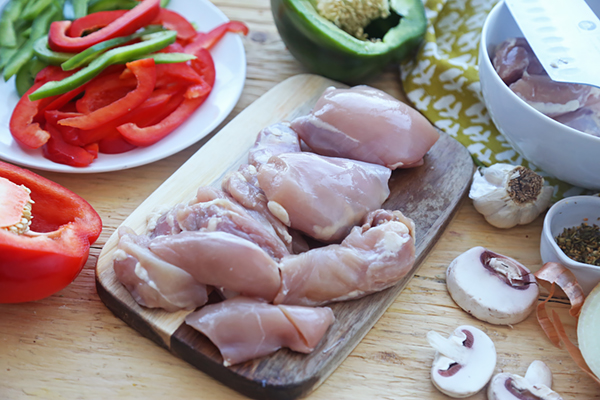 Raw chicken thighs on a cutting board with sliced bell peppers, mushrooms, and garlic