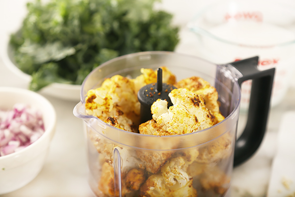 Roasted curry-coated cauliflower loaded into a food processor with diced red onion and kale nearby