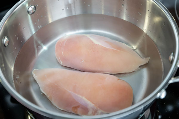 Two raw chicken breasts submerged in water inside a stainless steel pot