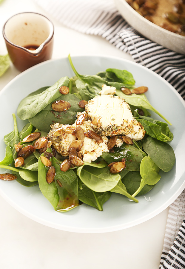 Baby spinach topped with baked goat cheese and balsamic butter pumpkin seeds in a white bowl